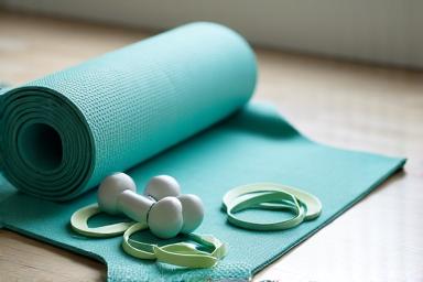 A collection of modern home fitness equipment, including a yoga mat, resistance bands, light dumbbells, and a foam roller, on a clean studio floor.
