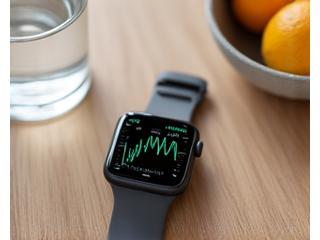 A smartwatch showing a detailed heart rate graph on its screen, lying on a wooden table with a blurred background of a healthy breakfast.